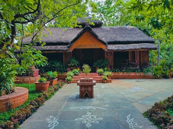 A charming rustic house with a thatched roof is surrounded by lush greenery. There are various potted plants and traditional floral patterns drawn on the stone pathway leading to the front door.