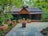 A charming rustic house with a thatched roof is surrounded by lush greenery. There are various potted plants and traditional floral patterns drawn on the stone pathway leading to the front door.