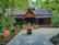 A charming rustic house with a thatched roof is surrounded by lush greenery. There are various potted plants and traditional floral patterns drawn on the stone pathway leading to the front door.