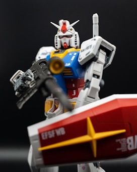 A detailed and well-crafted Gundam model figure stands against a dark background. The figure features a predominantly white and blue color scheme with red and yellow accents, holding a black gun in one hand and a red shield in the other. The model has intricate details, including various panels, markings, and a distinct V-shaped antenna on its head.