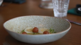 A shallow bowl with a light speckled texture contains a gourmet dish. There are round brown objects resembling small fruits or dumplings on a bed of sauce, accompanied by a thinly sliced white item. Green microgreens add a fresh touch. A glass is blurred in the background, placed on a wooden table.