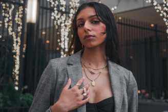A young woman wearing layered delicate necklaces and bold earrings, styled against a city backdrop.
