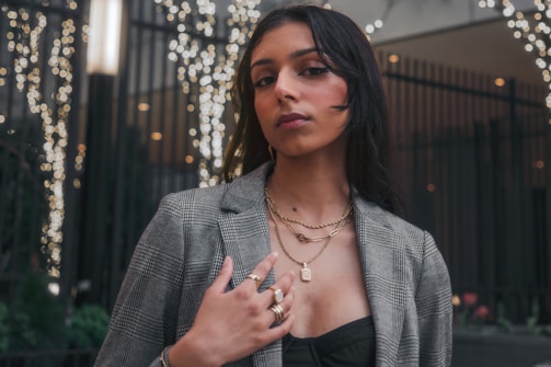 A person stands confidently in a stylish checkered blazer, layered gold necklaces, rings, and a black top. The background is decorated with blurred lights and dark railings, creating a festive or city night ambiance.