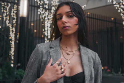 A young woman wearing layered delicate necklaces and bold earrings, styled against a city backdrop.