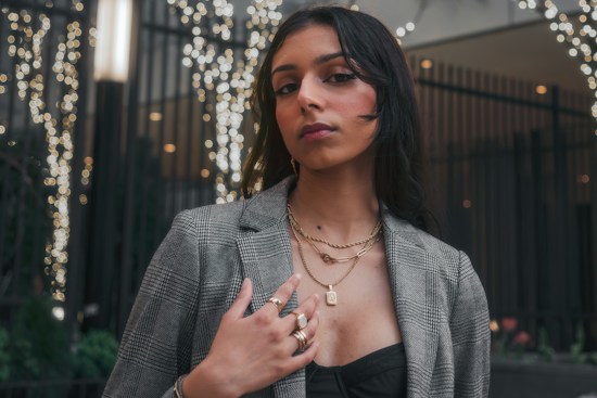 A person stands confidently in a stylish checkered blazer, layered gold necklaces, rings, and a black top. The background is decorated with blurred lights and dark railings, creating a festive or city night ambiance.