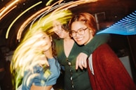 Three friends are embracing each other and smiling in a lively, energetic environment. The lighting is vibrant with streaks of swirling colors creating a dynamic and joyful atmosphere.