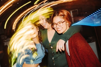 Three friends are embracing each other and smiling in a lively, energetic environment. The lighting is vibrant with streaks of swirling colors creating a dynamic and joyful atmosphere.