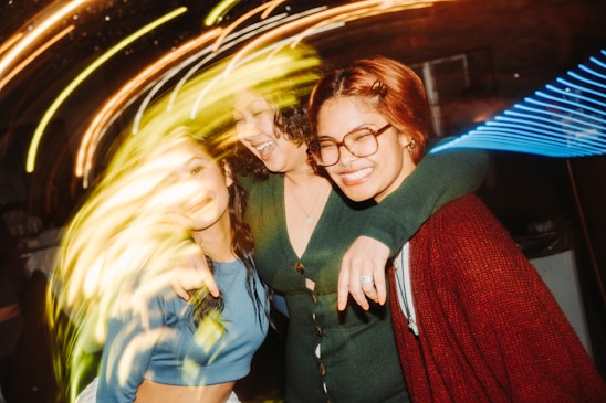 Three friends are embracing each other and smiling in a lively, energetic environment. The lighting is vibrant with streaks of swirling colors creating a dynamic and joyful atmosphere.