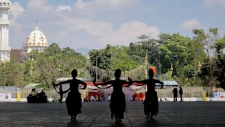 Three silhouetted dancers perform in front of a backdrop featuring lush greenery and a distinctive dome building with a golden pattern. The scene appears to be outdoors with a clear sky.