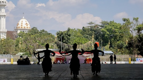 Three silhouetted dancers perform in front of a backdrop featuring lush greenery and a distinctive dome building with a golden pattern. The scene appears to be outdoors with a clear sky.
