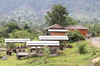 A rural setting with wooden structures, including a house and several gazebo-like shelters, surrounded by lush greenery and mountainous terrain in the background. People are seen sitting or gathered under the shelters, indicating a social or recreational setting.