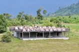 A group of people stands on a large wooden deck built above an open concrete structure. The deck is equipped with several dome-shaped tents or canopies, surrounded by lush green landscape. The background features a range of rolling hills or mountains, covered with dense forest.