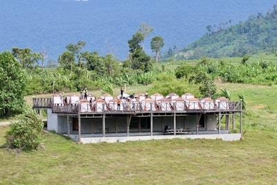 A group of people stands on a large wooden deck built above an open concrete structure. The deck is equipped with several dome-shaped tents or canopies, surrounded by lush green landscape. The background features a range of rolling hills or mountains, covered with dense forest.