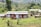 A collection of small cabins with wooden decks is set amidst a lush green landscape. The area is surrounded by a variety of trees and plants, and the ground is covered in vibrant grass. In the background, rolling hills are visible, adding depth to the scenery.