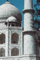 A close-up view of a large, intricately designed marble building with a prominent dome and minaret. The structure features ornate arches and detailed carvings, showcasing impressive craftsmanship. Sparse foliage is visible, adding contrast to the solid, structured architecture.