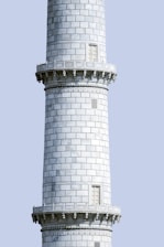 A tall, cylindrical stone tower with intricately carved details and decorative patterns. The surface is composed of uniform rectangular stone slabs, interrupted by narrow windows. The tower features horizontal bands with ornamental designs just below the ledges.