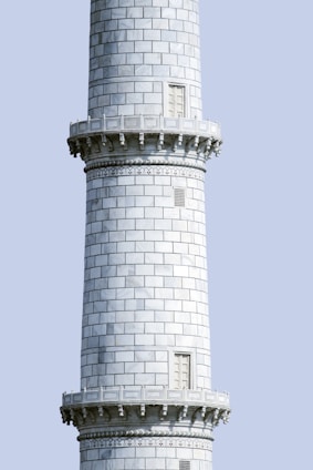 A tall, cylindrical stone tower with intricately carved details and decorative patterns. The surface is composed of uniform rectangular stone slabs, interrupted by narrow windows. The tower features horizontal bands with ornamental designs just below the ledges.