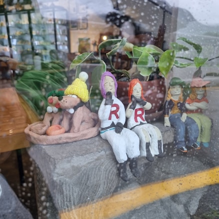 Clay figurines are sitting on a display next to a potted plant inside a shop. The figures include dogs wearing knitted hats and scarves, and characters dressed in white with red 'R' letters on their chests. The scene is viewed from outside through a window with rain droplets.