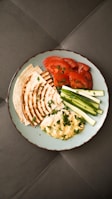 A plate containing slices of grilled flatbread, fresh tomato slices, cucumber sticks, and a serving of hummus garnished with chopped parsley. The plate is set against a dark, textured surface.