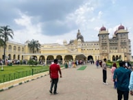 A large, ornate palace stands under a cloudy sky. The building features intricate architectural details such as domes and arches. A group of people are gathered in front of the structure, walking along a pathway lined with green grass and decorative fencing. Several palm trees provide additional greenery around the area.