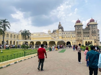 A large, ornate palace stands under a cloudy sky. The building features intricate architectural details such as domes and arches. A group of people are gathered in front of the structure, walking along a pathway lined with green grass and decorative fencing. Several palm trees provide additional greenery around the area.
