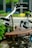 The image focuses on a vintage bicycle seat with a brown leather surface and metal springs, prominently featuring the brand name 'Lepper'. The background is softly blurred, showing hints of greenery and an urban setting.