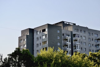 A modern, multi-story apartment building with a flat roof and several balconies is surrounded by lush green trees. The building is predominantly grey with multiple windows, and the sky appears clear and blue.