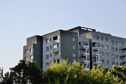 A modern, multi-story apartment building with a flat roof and several balconies is surrounded by lush green trees. The building is predominantly grey with multiple windows, and the sky appears clear and blue.