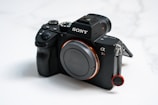 A black Sony camera with a textured grip and prominent branding. The model is the Alpha 7R with visible buttons and a lens cap protecting the front. The background is softly blurred, drawing attention to the device.