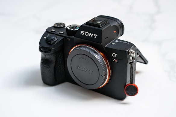 A black Sony camera with a textured grip and prominent branding. The model is the Alpha 7R with visible buttons and a lens cap protecting the front. The background is softly blurred, drawing attention to the device.