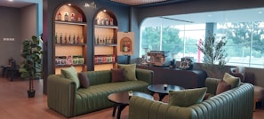 A modern lounge interior with green upholstered seating arranged around circular black coffee tables. Shelves on the back wall display a variety of liquor bottles and boxes. There is a coffee machine setup with accessories on the right side of the shelves. Large windows on the right allow natural light to enter, providing a view of greenery outside. There are also a couple of plants placed around the room for decoration.