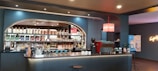 A modern and stylish coffee bar features a well-stocked back wall with a variety of bottles and packaged coffee products on shelves. The counter is sleek with a coffee machine, cups, and barista tools. A neon sign adds a touch of vibrant color on the wall.