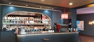 A modern and stylish coffee bar features a well-stocked back wall with a variety of bottles and packaged coffee products on shelves. The counter is sleek with a coffee machine, cups, and barista tools. A neon sign adds a touch of vibrant color on the wall.