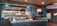 A modern and stylish coffee bar features a well-stocked back wall with a variety of bottles and packaged coffee products on shelves. The counter is sleek with a coffee machine, cups, and barista tools. A neon sign adds a touch of vibrant color on the wall.