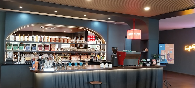 A modern and stylish coffee bar features a well-stocked back wall with a variety of bottles and packaged coffee products on shelves. The counter is sleek with a coffee machine, cups, and barista tools. A neon sign adds a touch of vibrant color on the wall.