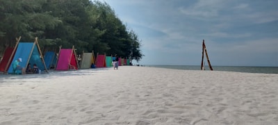 A serene beach setting with white sand stretching towards the horizon. There are colorful tents or cabanas lined up on the left side, with a few people sitting under them. The right side opens to a vast ocean under a partly cloudy sky. Trees are visible near the tents, providing some shade.