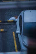A white car is parked in a wet parking lot, with visible rain droplets creating a textured effect on the surface. The car is positioned beside yellow and black striped barriers. A white bag or sack is placed on the ground nearby.
