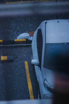 A white car is parked in a wet parking lot, with visible rain droplets creating a textured effect on the surface. The car is positioned beside yellow and black striped barriers. A white bag or sack is placed on the ground nearby.