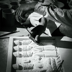 A craftsman is engaged in detailed carving work on a stone slab, depicting intricate human figures. The hands holding tools are in focus, demonstrating precision and skill.