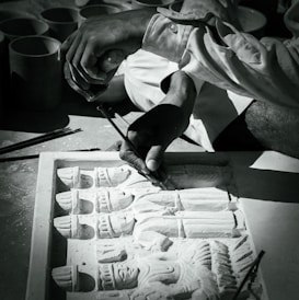 A craftsman is engaged in detailed carving work on a stone slab, depicting intricate human figures. The hands holding tools are in focus, demonstrating precision and skill.