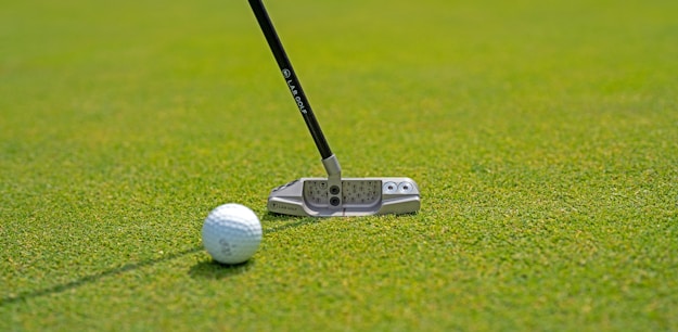 A close-up view of a golf club putter next to a golf ball on a well-maintained green. The focus is on the detail of the putter and the texture of the grass.