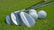 Several golf clubs with shiny metallic surfaces are laid on a lush green grass surface next to a few white golf balls. The focus captures the fine details of the club heads and the dimpled texture of the golf balls.