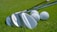 Several golf clubs with shiny metallic surfaces are laid on a lush green grass surface next to a few white golf balls. The focus captures the fine details of the club heads and the dimpled texture of the golf balls.
