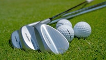 Several golf clubs with shiny metallic surfaces are laid on a lush green grass surface next to a few white golf balls. The focus captures the fine details of the club heads and the dimpled texture of the golf balls.