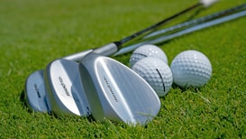 Several golf clubs with shiny metallic surfaces are laid on a lush green grass surface next to a few white golf balls. The focus captures the fine details of the club heads and the dimpled texture of the golf balls.