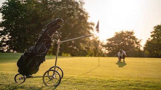A golf course with a bag of golf clubs on a wheeled trolley in the foreground. The surface is well-maintained grass, and there is a flag on the green. In the distance, a person crouches, possibly lining up a shot. The scene is lit by warm sunlight, creating a serene environment.