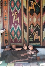 A brightly colored woven tapestry with geometric and horse motifs covers the wall. Below it, a table draped with a dark fabric displays an assortment of clay pottery, including vases and bowls.