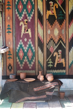 A brightly colored woven tapestry with geometric and horse motifs covers the wall. Below it, a table draped with a dark fabric displays an assortment of clay pottery, including vases and bowls.