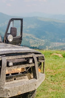 A rugged off-road vehicle is partially visible, with its front section shown against a backdrop of rolling green hills under a light blue sky. The vehicle, covered in dirt, suggests recent usage on rough terrain. Its door is open, and its headlights are prominently featured.