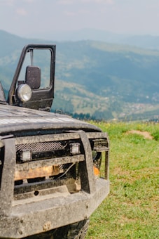 A rugged off-road vehicle is partially visible, with its front section shown against a backdrop of rolling green hills under a light blue sky. The vehicle, covered in dirt, suggests recent usage on rough terrain. Its door is open, and its headlights are prominently featured.